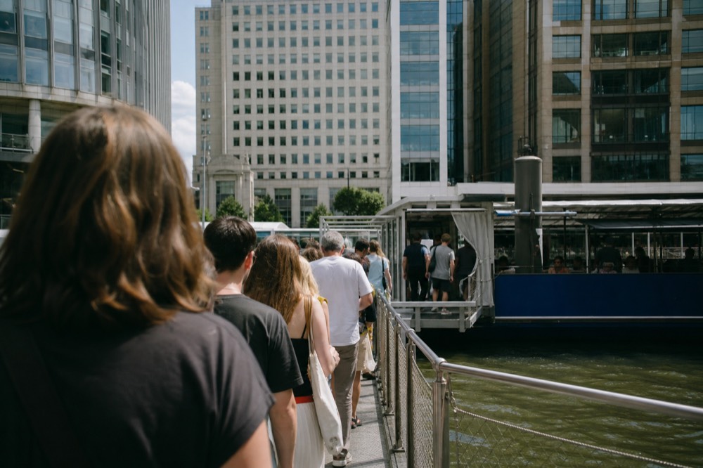 A line of people waiting along a floating pier rail in bright summer light, a boat's blue hull and open gangway ahead of them