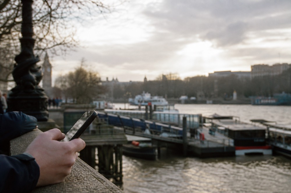 A hand holding a phone at a riverside railing on an overcast afternoon, a pier and moored boats out of focus beyond
