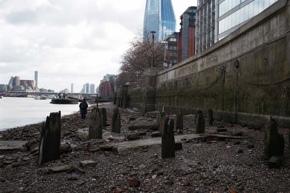 Exposed Thames foreshore at low tide, dark shingle scattered with brick fragments and worn timber piles, embankment wall rising behind under flat light