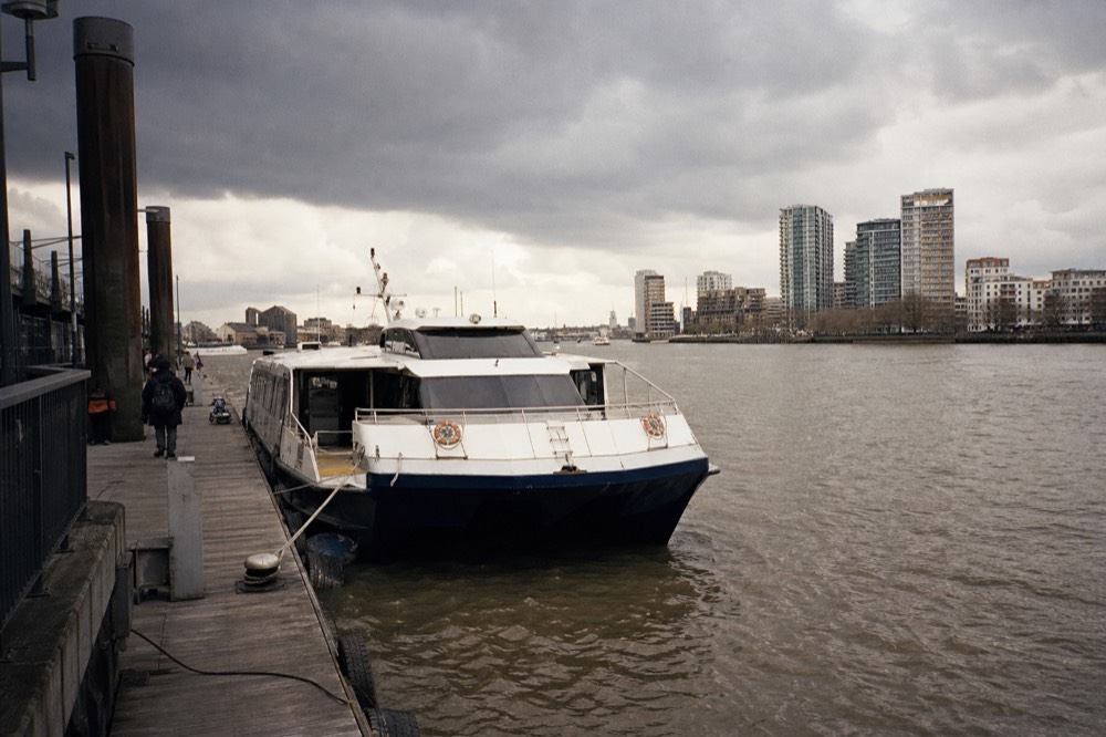 A twin-hulled commuter catamaran tied alongside a floating pontoon pier with its boarding door open and the river running past