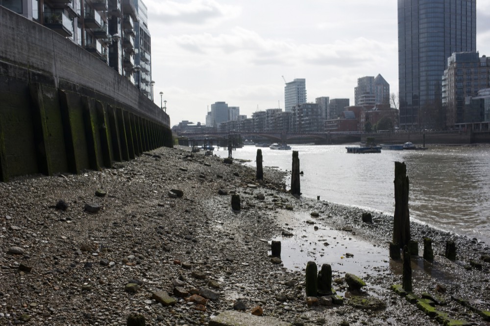 Wide exposed shingle foreshore at low tide below a curved embankment wall, rotted timber piles standing in shallow pools and the river narrowed to a channel beyond