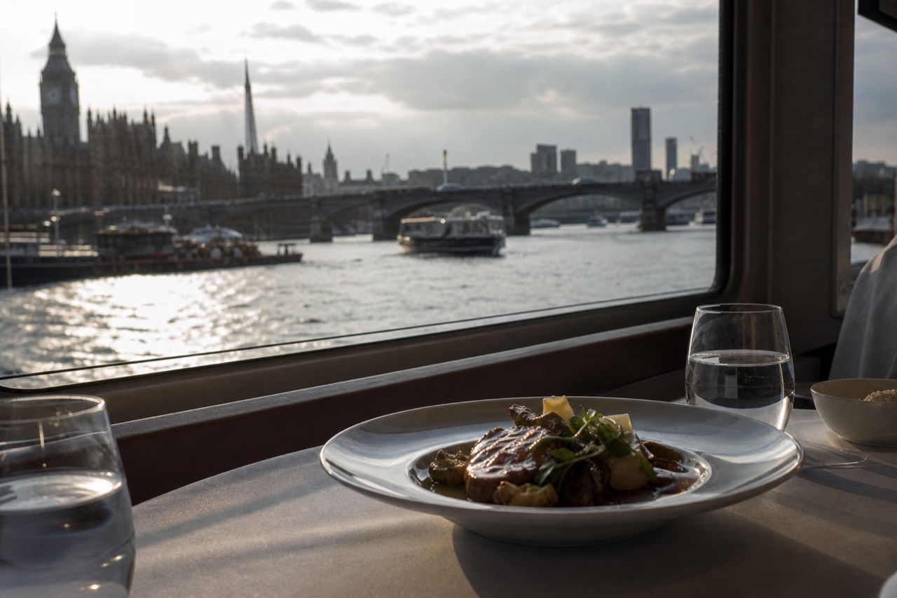 A laid table beside a boat window in daylight with a plated main course and water glasses, the river and a bridge outside