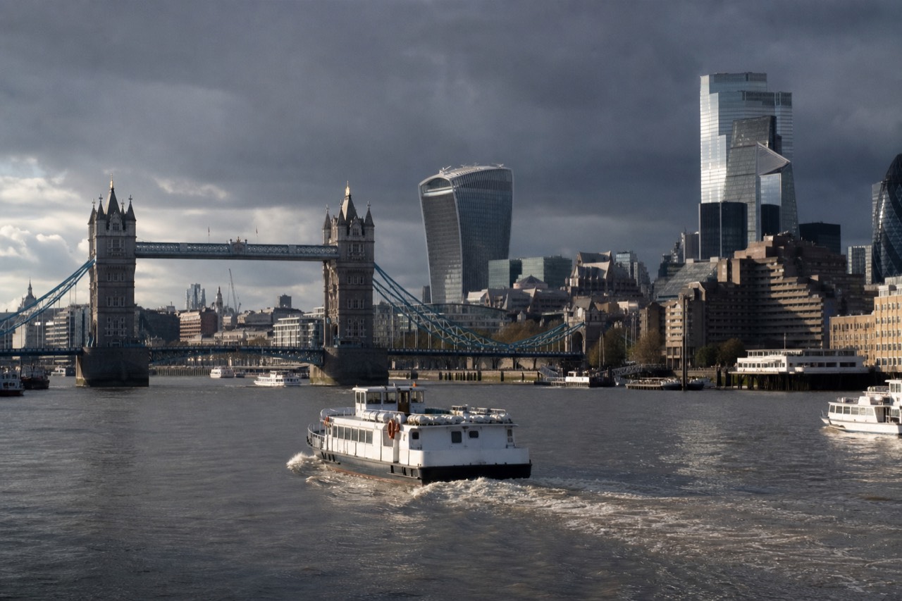 A passenger boat mid-river on a bright morning with Tower Bridge behind it and the City towers rising along the north bank