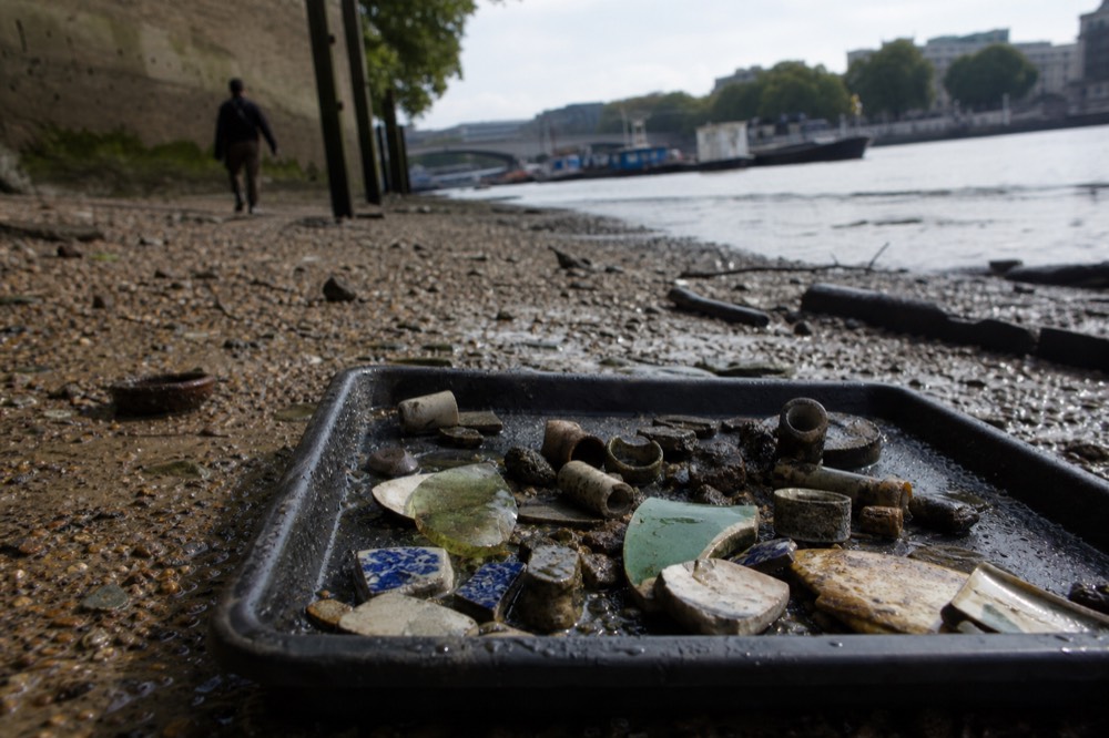 A shallow tray of small river-worn objects on wet gravel, clay pipe bowls, corroded pins, fragments of glazed pottery and a green-tinted glass shard