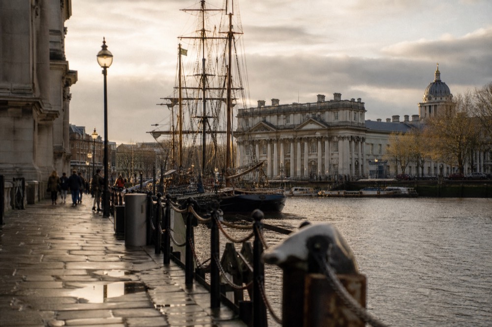 A riverside pier at Greenwich with tall ship masts rising behind it and classical stone buildings along the waterfront