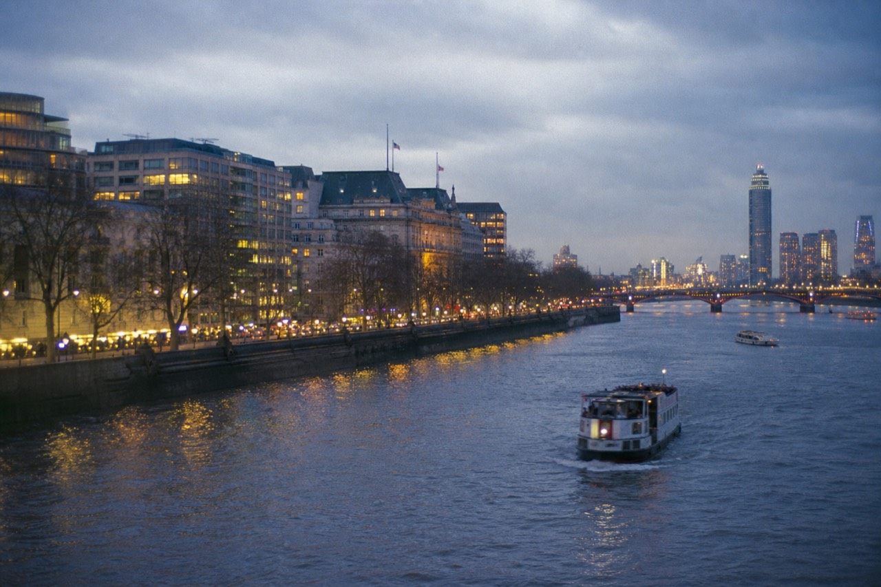 Wide view along the Thames at dusk with the lit riverfront curving away, bridge arches receding into blue haze and reflections breaking on the water