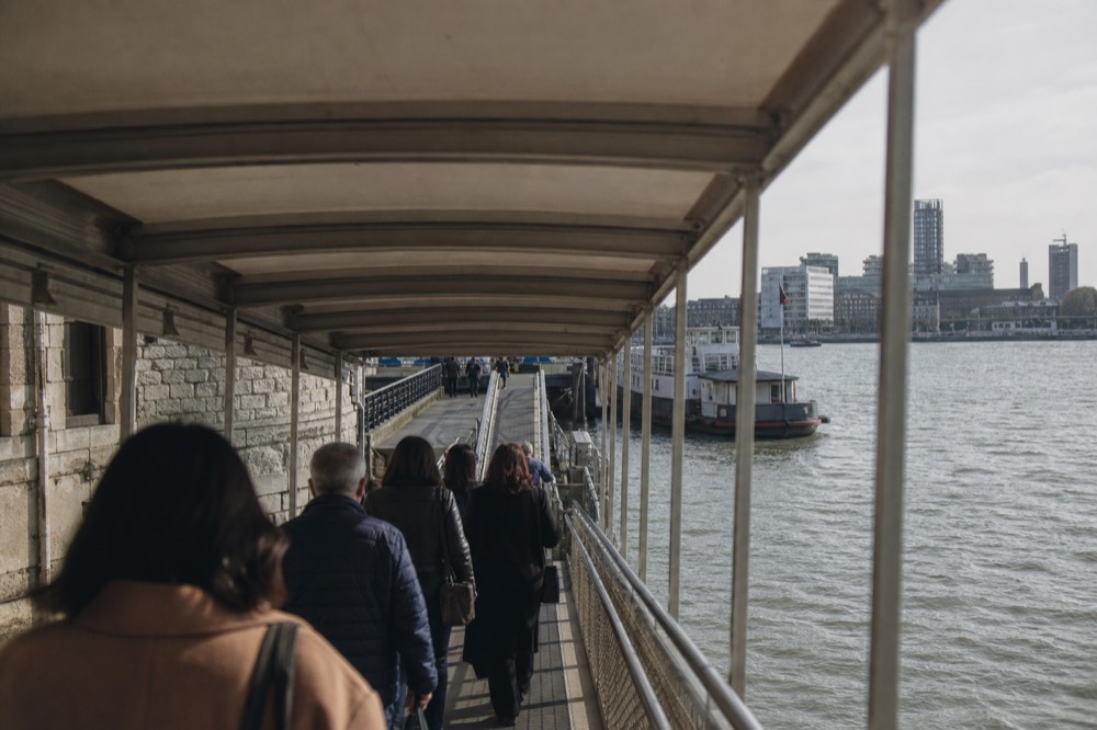 Passengers walking down a covered gangway to a floating pier beside the stone walls of a riverside fortress on a bright afternoon