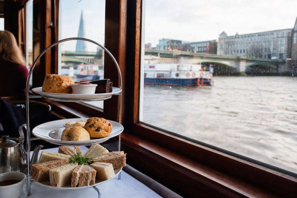A tiered cake stand of sandwiches and scones on a white cloth beside a boat window, the river and a bridge visible outside