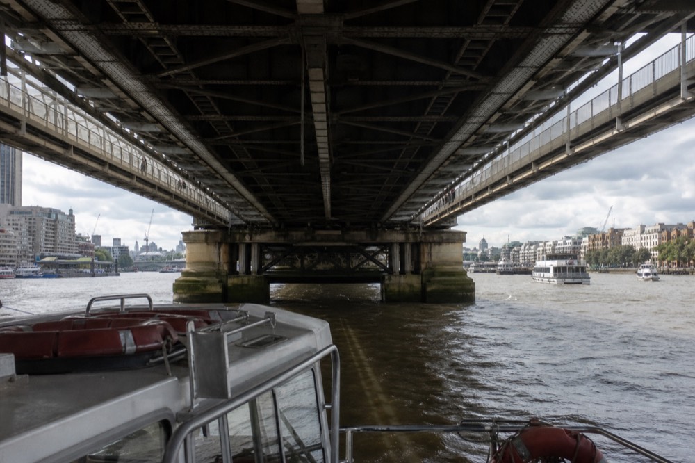 The underside of a steel railway bridge with pedestrian decks either side, seen from a boat passing beneath on a bright afternoon