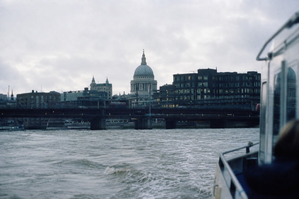 A pale stone cathedral dome rising above dark office blocks on the north bank of a wide river, seen low from a passing boat