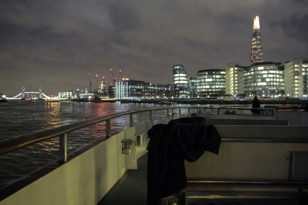 An empty open boat deck at night with a coat draped over a bench and the rail wet, city lights blurred beyond