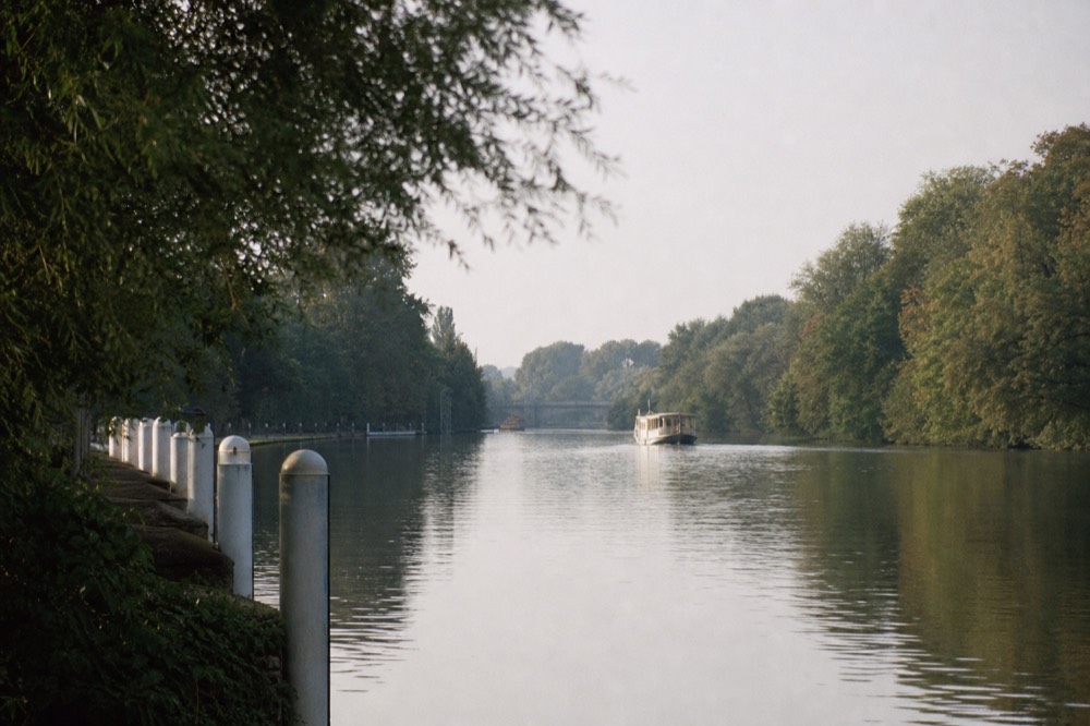 A long straight reach of river between densely wooded banks in late afternoon light, white marker posts along one side and flat still water