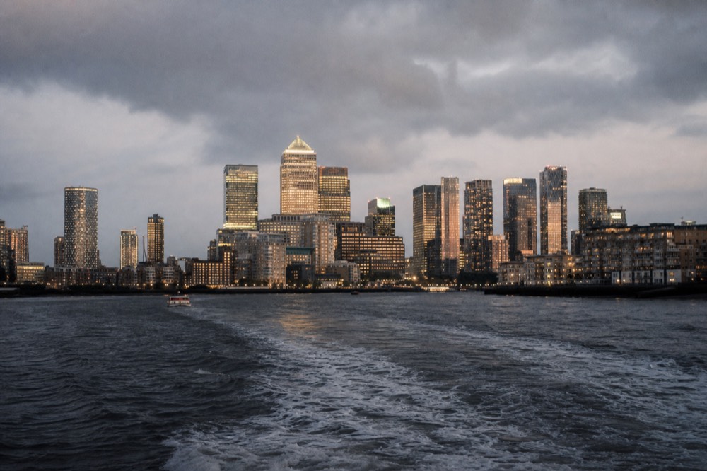 The lit towers of Canary Wharf seen low across dark water at night, their reflections broken by a boat wake