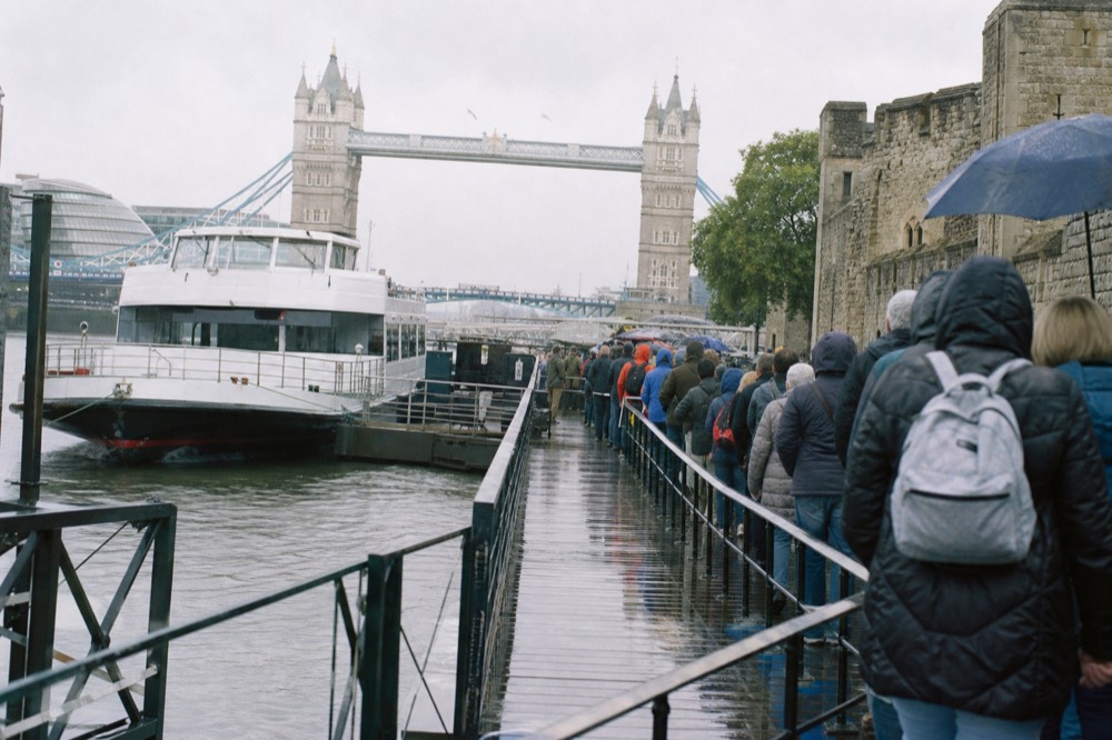 A floating pier beside a stone riverside fortress at midday, a white cruise boat alongside and a queue of passengers on the pontoon
