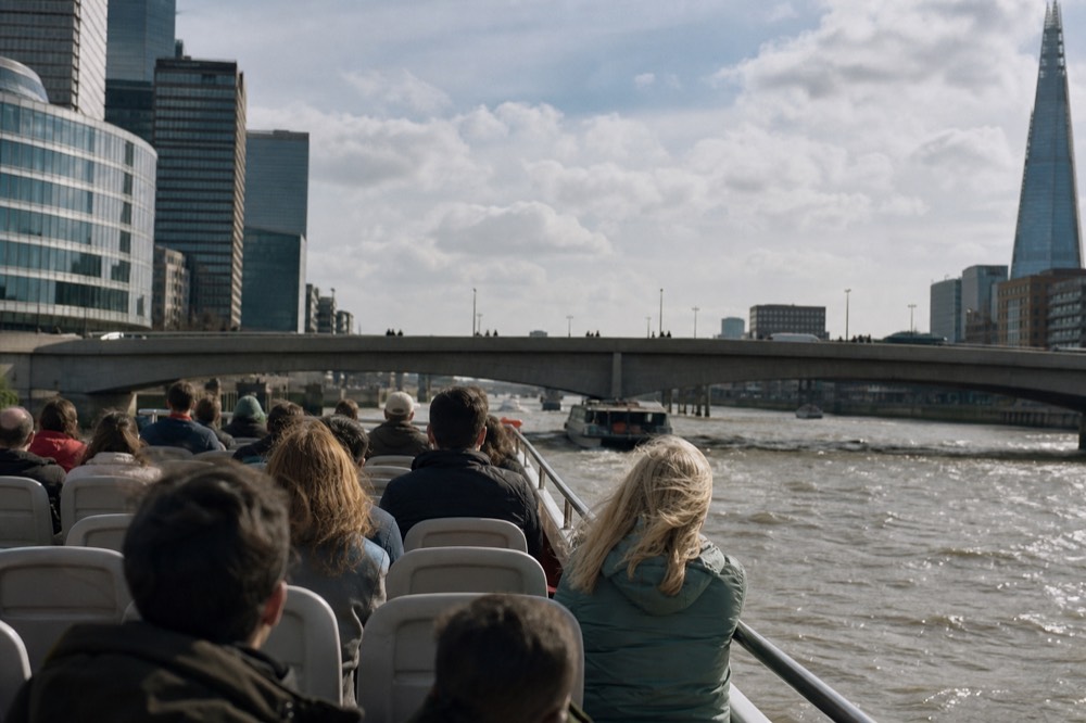 Passengers seated along the open upper deck of a river boat on a breezy bright day, a bridge span approaching ahead