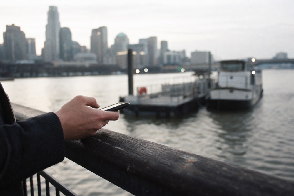 A hand holding a phone against a river railing on a grey afternoon, a pier and moored boat out of focus beyond