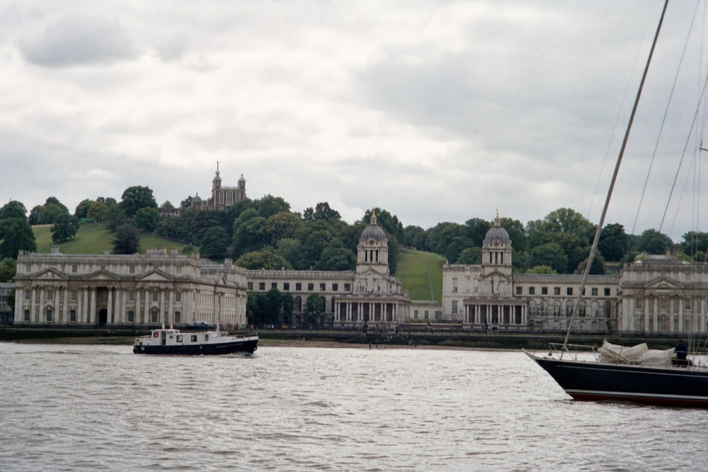 The white colonnaded buildings of a riverside naval college seen from mid-river, green hill and observatory behind