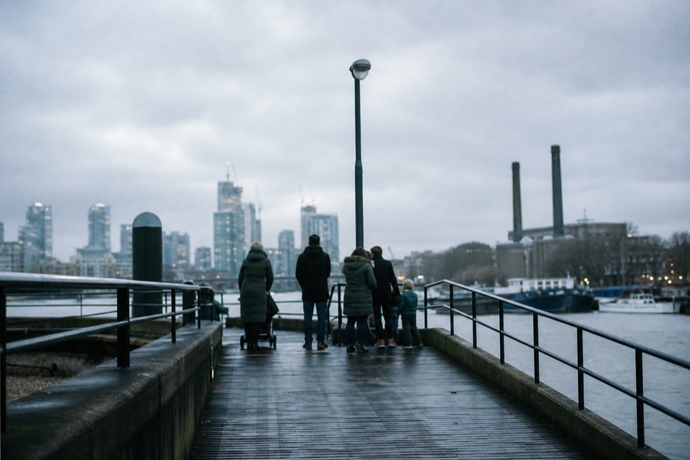 A pushchair and a small group waiting on a sloping pier ramp beside the river railings on an overcast day, seen from behind