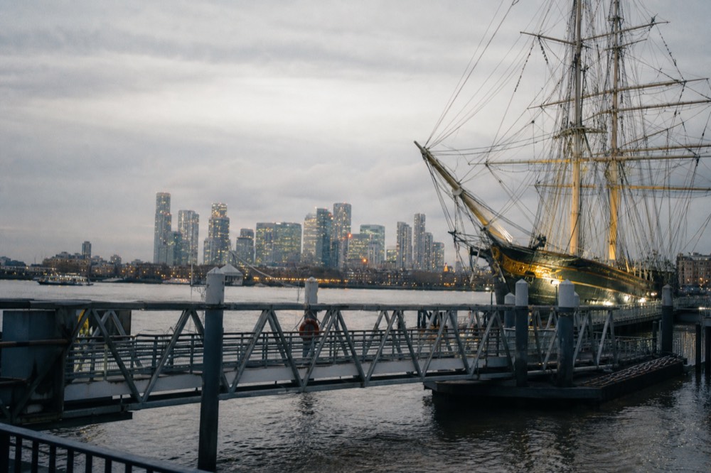 A river pier at Greenwich with the tall masts and gilded hull of a preserved clipper ship rising behind the pontoon on an overcast afternoon